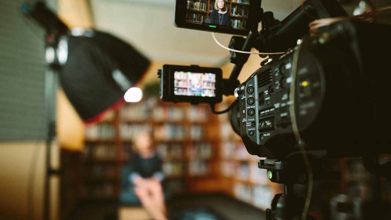 A camera set up in the foreground interviewing a woman who is sitting in a blurred background on a chair in a library