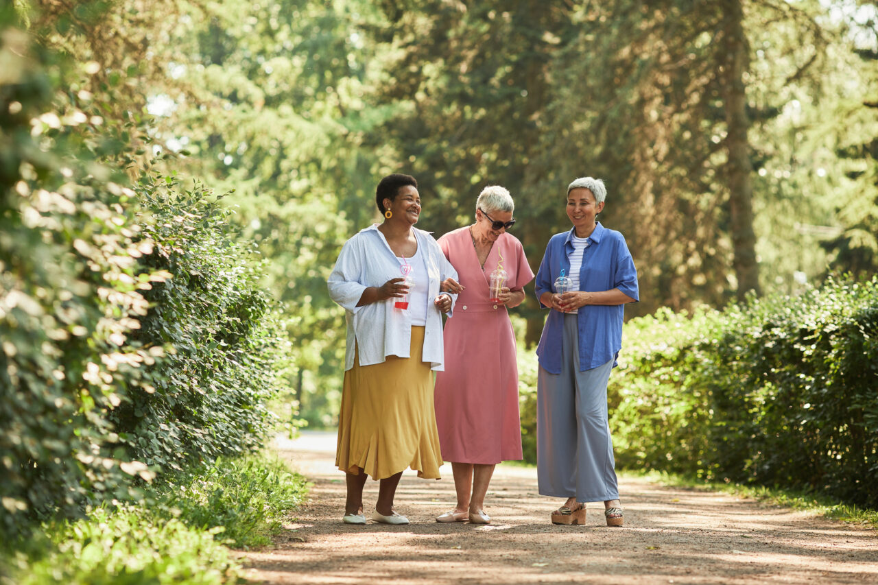 Three older women walking down a path while holding drinks and laughing