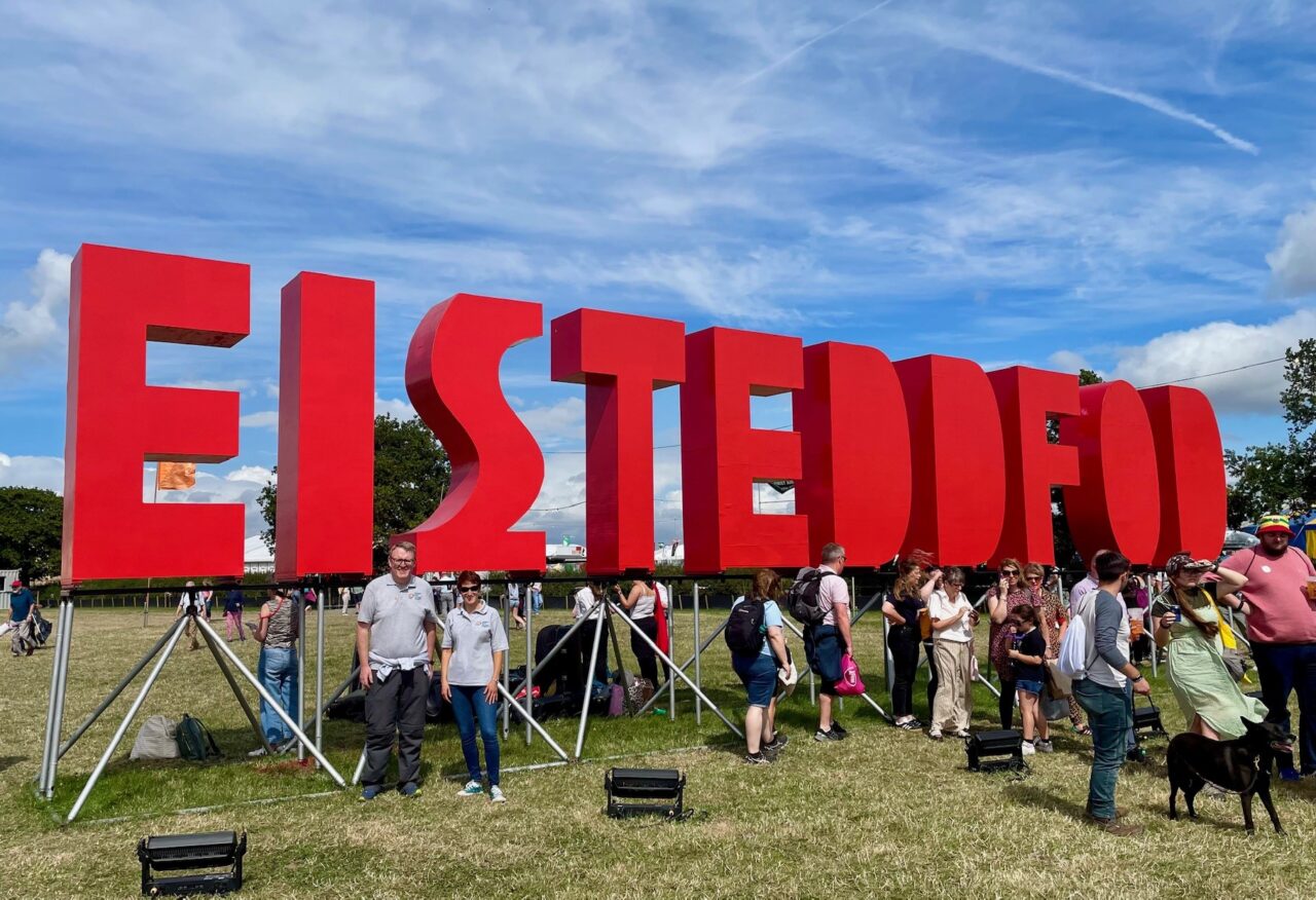 Delwedd o Rhian Bowen-Davies a'i chydweithiwr o flaen yr arwydd coch mawr 'EISTEDDFOD'.