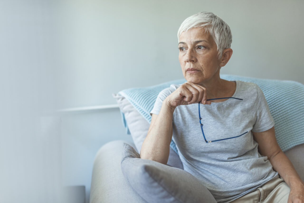 Delwedd o fenyw hŷn â gwallt gwyn byr, yn eistedd ar soffa, yn edrych allan o'r ffenestr. // Image of an older woman with short white hair, sat on a sofa, looking out of the window.