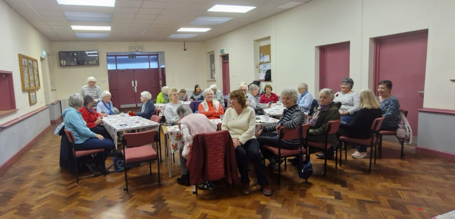 Delwedd o grŵp o bobl hŷn yn eistedd o amgylch bwrdd mawr yng Nghlwb Cinio Carew. // Image of a group of older people sat around a large table at Carew Lunch Club.