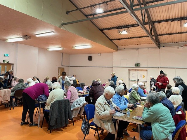 Eisteddodd grŵp mawr o bobl hŷn ar draws sawl bwrdd, yn gwneud cardiau Nadolig. // A large group of older people sat across several tables, making Christmas cards.