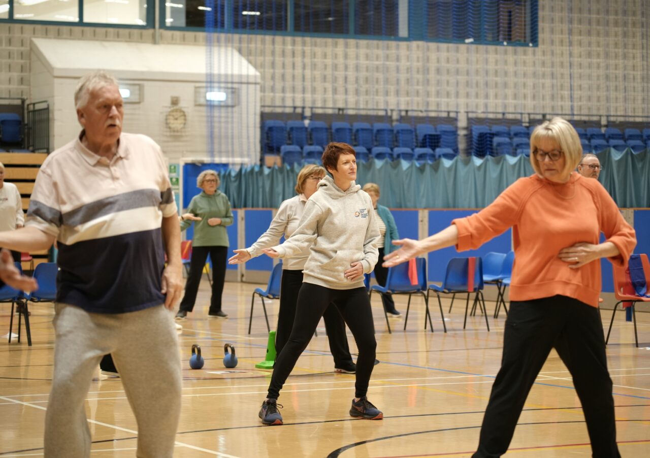 Comisiynydd Pobl Hŷn Cymru, Rhian Bowen-Davies (canol), yn y llun yn mynychu dosbarth ffitrwydd yng Nghanolfan Hamdden Halo. O'i chwmpas mae nifer o bobl hŷn sydd hefyd yn cymryd rhan yn y sesiwn. // Older People's Commissioner for Wales, Rhian Bowen-Davies (centre), pictured attending a fitness class at the Halo Leisure Centre. Around her are several older people who are also taking part in the session.