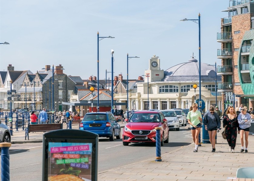 Delwedd o ganol tref Porthcawl, gyda cheir yn gyrru drwy'r brif stryd, a phobl yn cerdded ar y stryd fawr. // Image of Porthcawl town centre, with cars driving through the main street, and people walking on the high street.