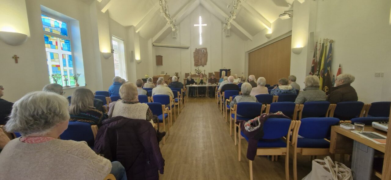 Delwedd o dros 20 o bobl hŷn yn eistedd mewn cyfarfod o U3A Torfaen, y tu mewn i neuadd eglwys. // Image of over 20 older people sat at a meeting of the Torfaen U3A, inside a church hall.