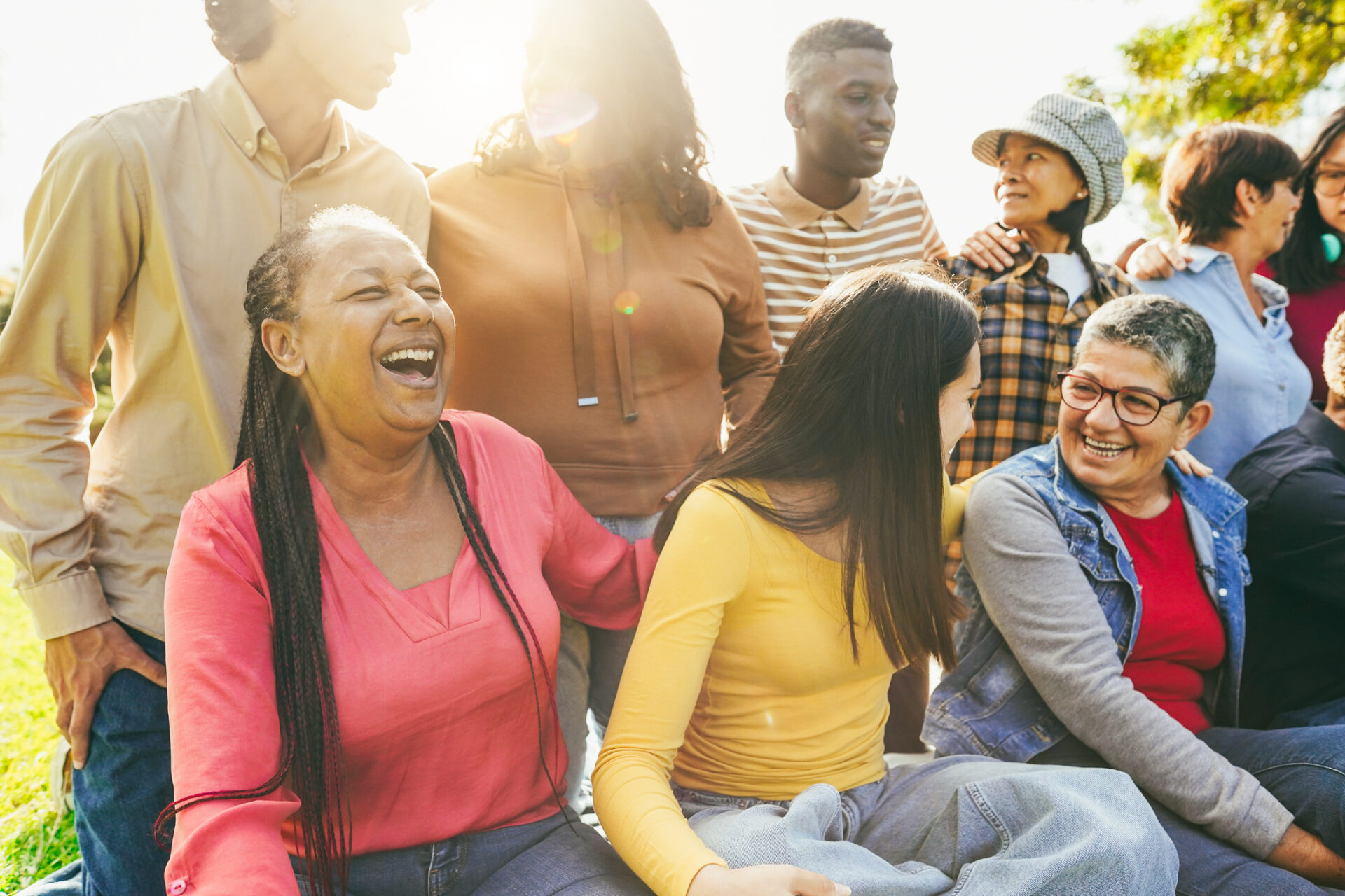 Delwedd o grŵp o oedolion o wahanol oedrannau, rhai yn eistedd ar y llawr a rhai yn sefyll, pob un yn gwenu ac yn chwerthin. / Image of a group of adults of a variety of ages, some sat on the floor and some standing up, all smiling and laughing.