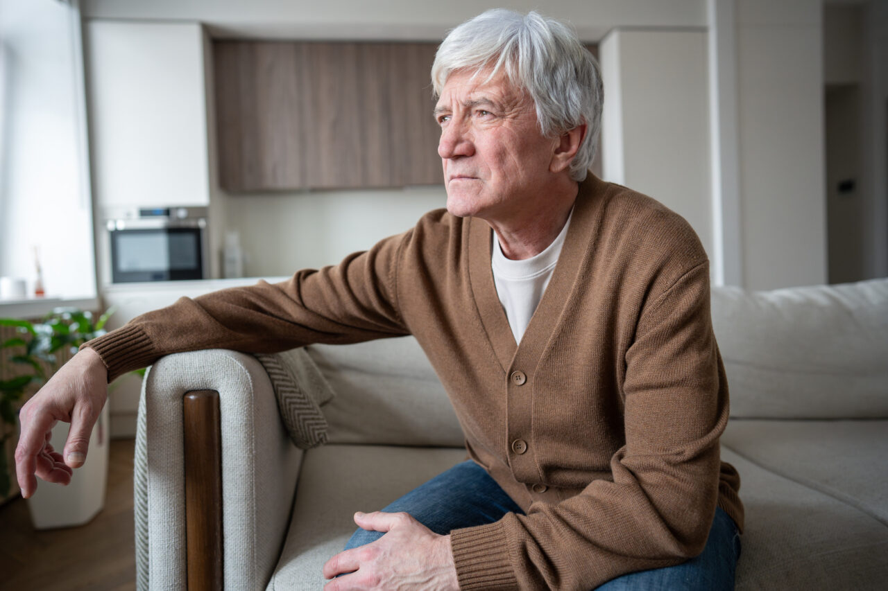 Eisteddodd dyn hŷn ar soffa mewn ystafell fyw, gan edrych allan ar draws yr ystafell. // Older man sat down on a sofa in a living room, looking out across the room.