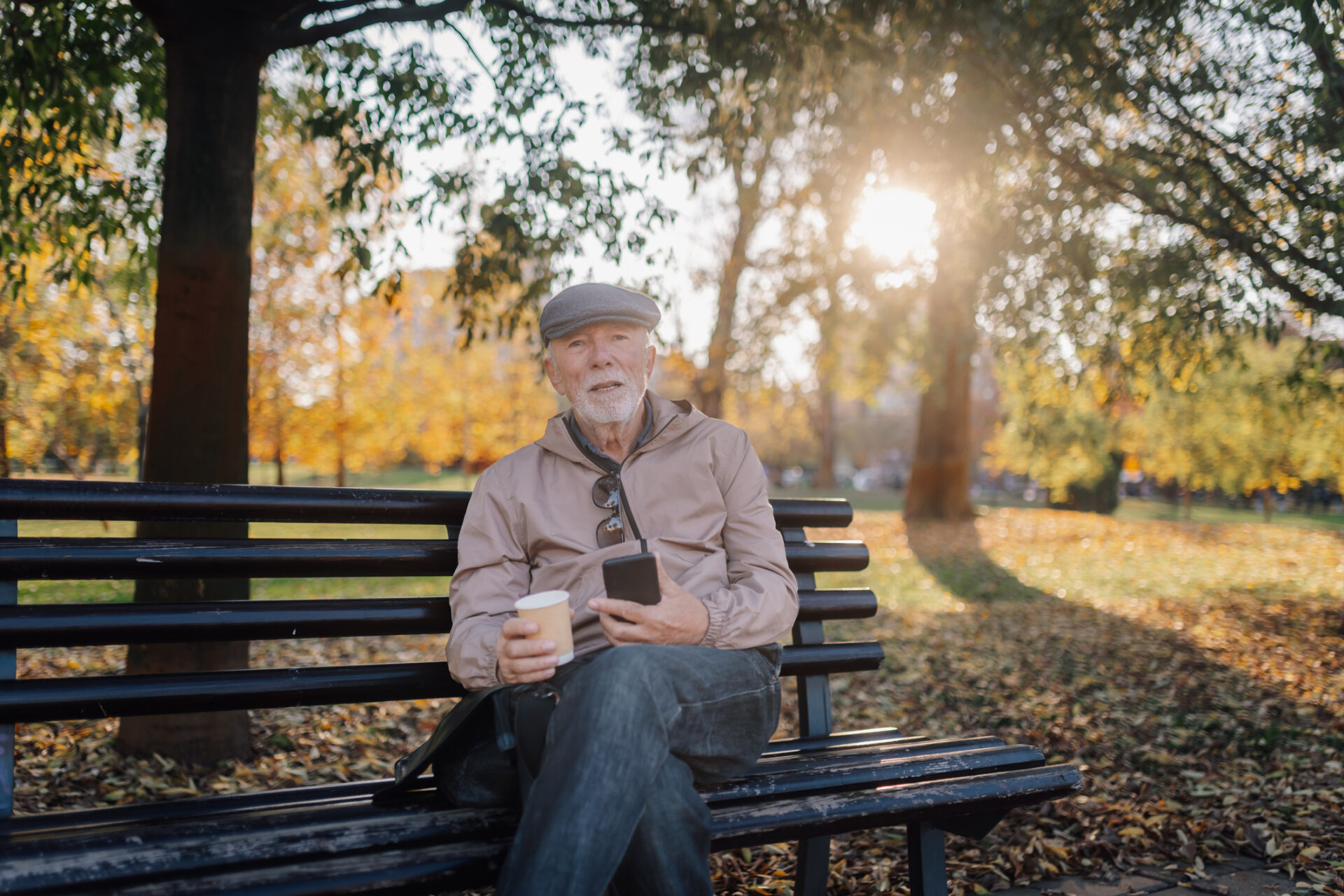 Dyn hŷn yn eistedd ar fainc parc, yn dal ffôn clyfar a choffi, yn mwynhau diwrnod tawel yn yr hydref yn yr awyr agored // Elder man sitting on a park bench, holding a smartphone and coffee, enjoying a tranquil autumn day outdoors