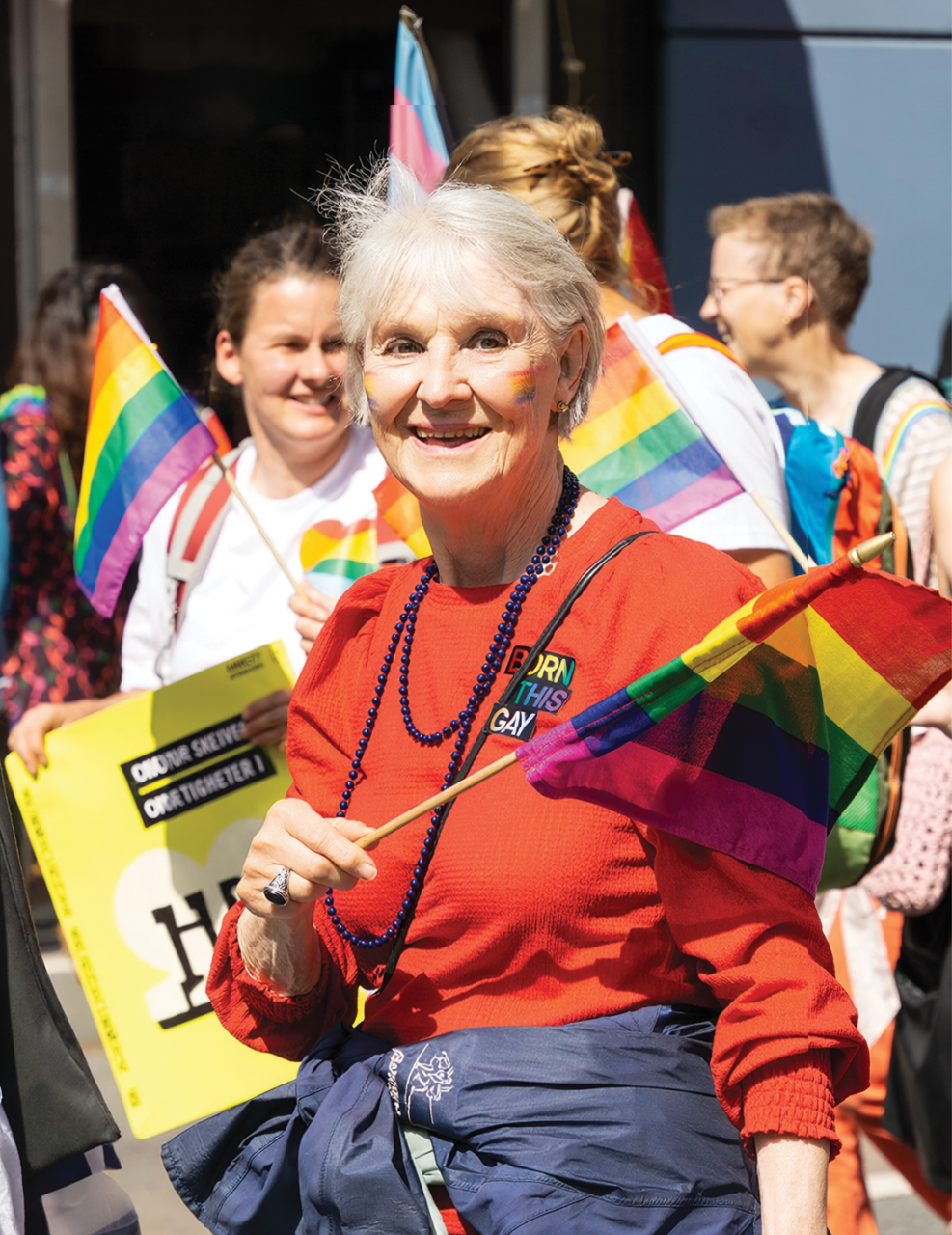 Photograph of a participant at a Pride event holding a rainbow flag. The participant is wearing a bright red top, blue bead necklaces and a jacket tied around the waist. Several other participants are visible in the background, some holding rainbow and transgender pride flags. One person is carrying a yellow sign. The image is brightly lit by sunlight and features prominent rainbow colours throughout.